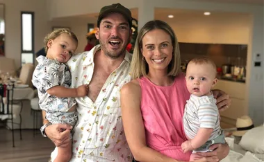 A smiling family of four in casual clothing indoors, holding their two young children.