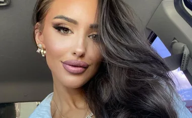 A woman with long dark hair poses in a car, wearing makeup and earrings.
