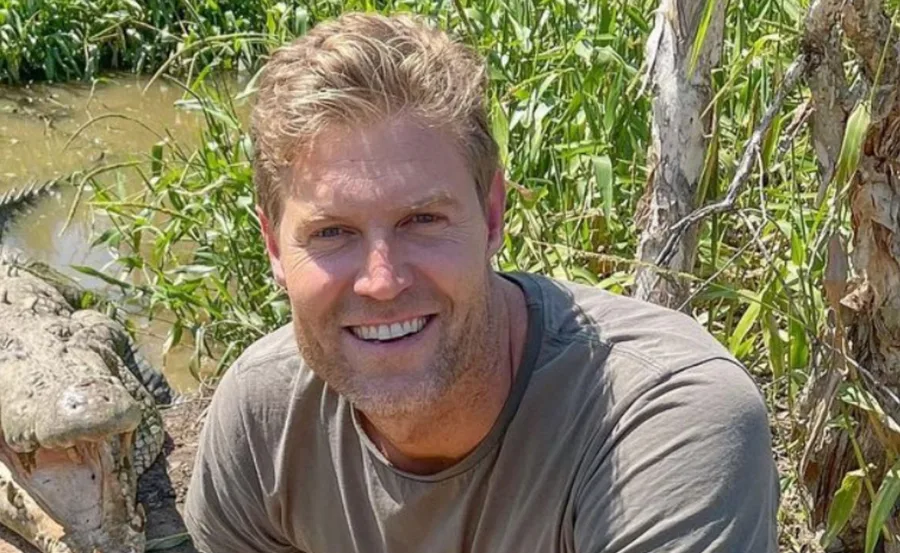 Man smiling near open-mouthed crocodile, surrounded by greenery.