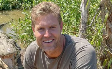 Man smiling near open-mouthed crocodile, surrounded by greenery.