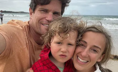 A family selfie on a beach with a man, woman, and child smiling at the camera, ocean waves in the background.