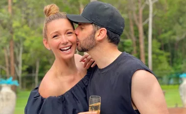 A man kisses a smiling woman's cheek in a garden, both looking joyful.