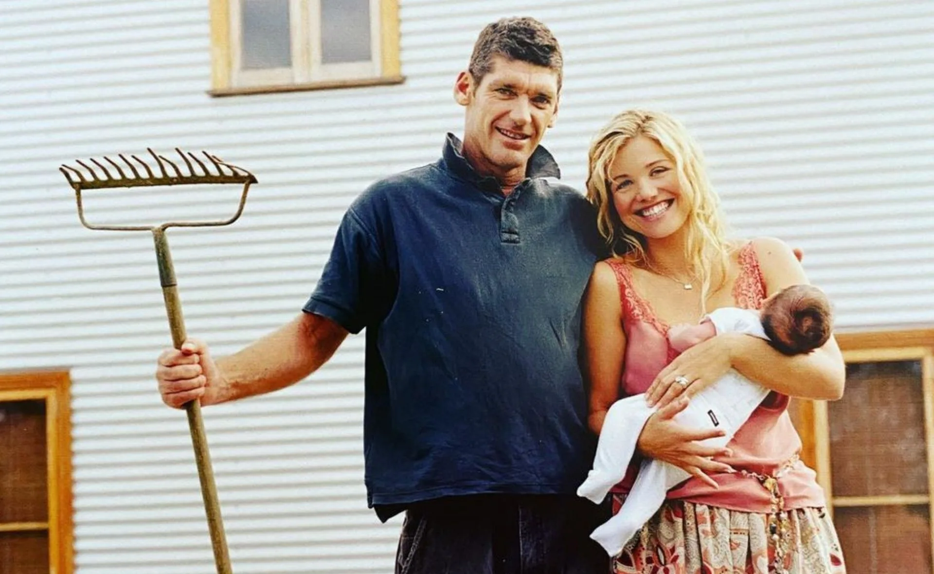 A couple with a baby, man holding a rake, smiling in front of a corrugated metal building.