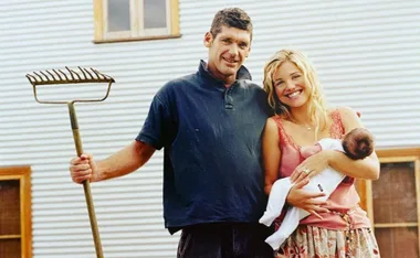 A couple with a baby, man holding a rake, smiling in front of a corrugated metal building.