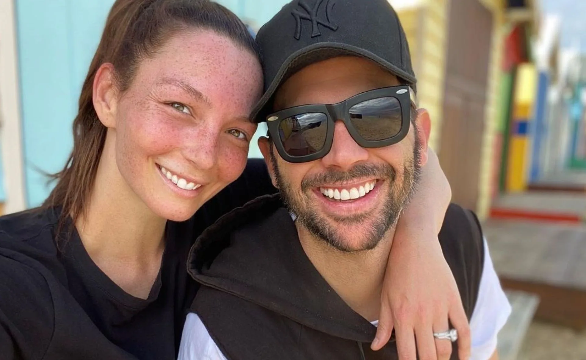 A smiling couple embraces outdoors; the man wears a cap and sunglasses.