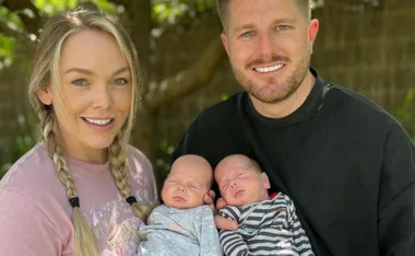 A couple smiles while holding their sleeping newborn twins outside on a sunny day.