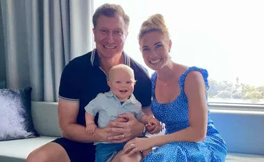 A smiling family of three sits together, with a man, woman, and baby on a sofa by a window.