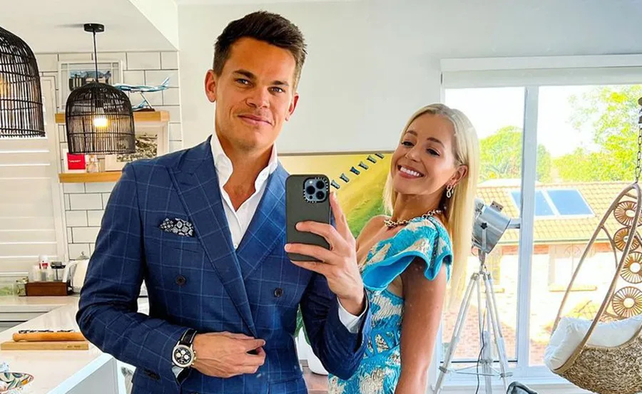 A man in a blue suit and a woman in a blue dress smile together, taking a mirror selfie in a modern kitchen.