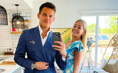 A man in a blue suit and a woman in a blue dress smile together, taking a mirror selfie in a modern kitchen.