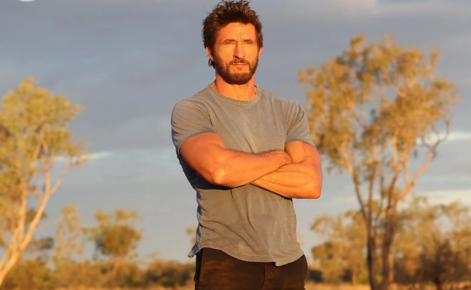 Man with crossed arms standing outdoors during sunset, trees in the background.