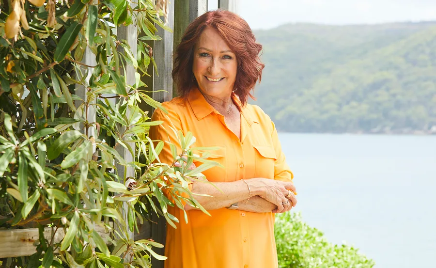 Woman in orange shirt smiling by a wooden fence with greenery, hills, and water in the background.
