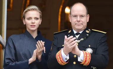 A couple in formal attire applauds from a balcony, both appearing serious and composed.