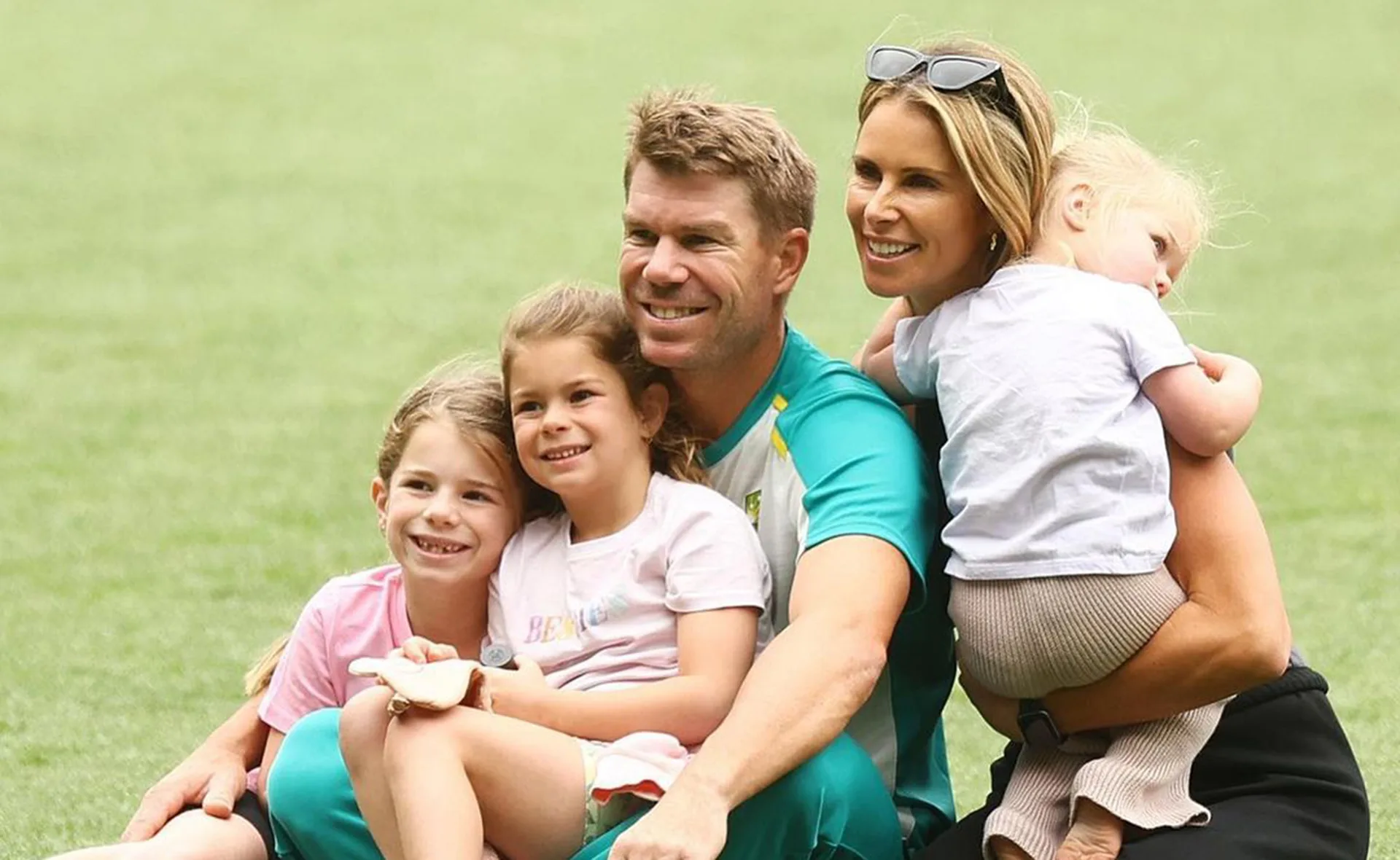 Family sitting on grass, smiling; parents with three young children in a cozy, affectionate pose.