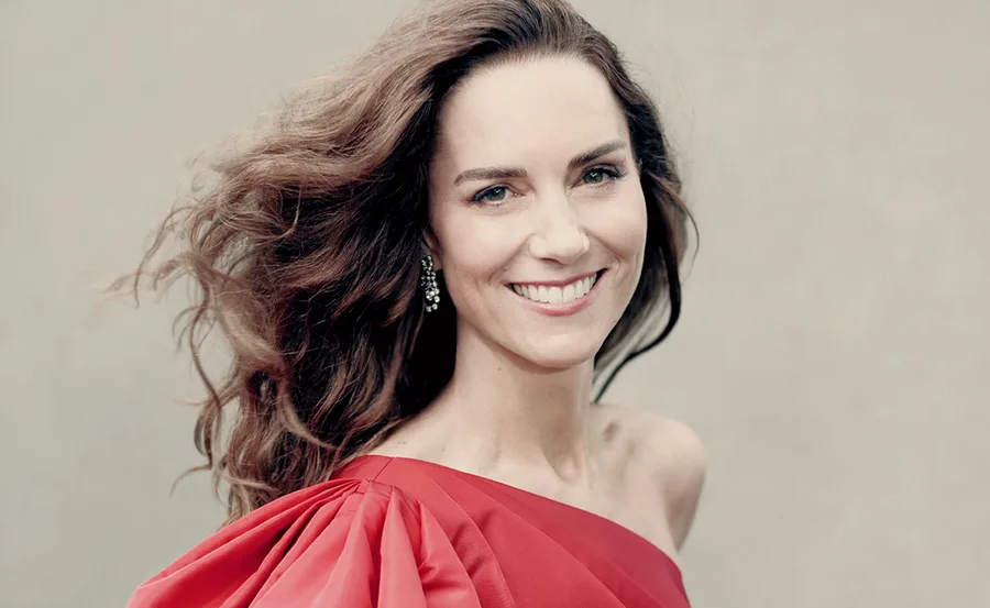A woman with long hair and earrings smiles in a red dress, posing against a plain background.