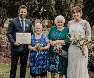 A couple poses with two elderly women at an outdoor wedding, holding flowers and a certificate.