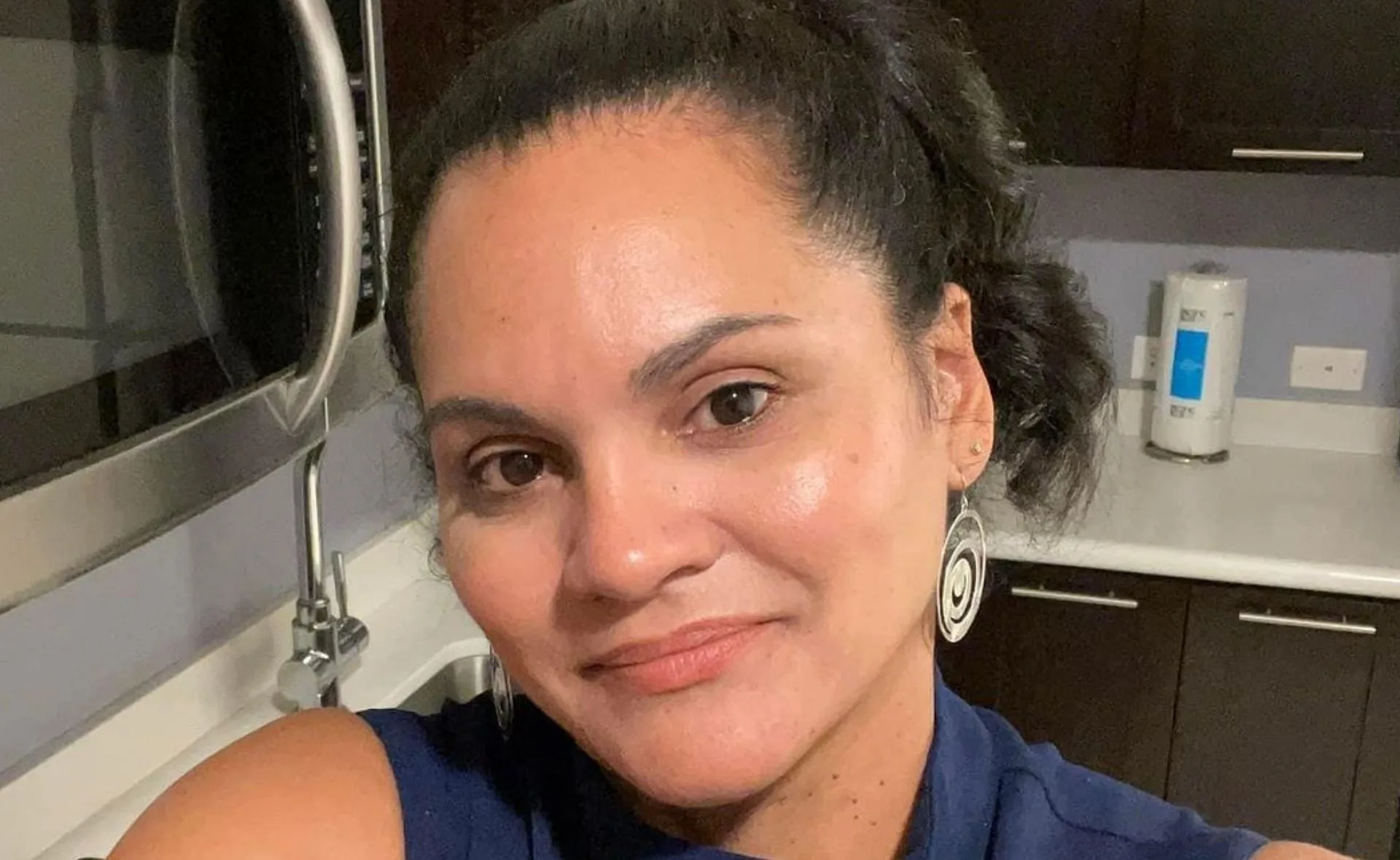 A woman with dark hair pulled back, wearing large earrings, takes a selfie in a kitchen with modern appliances.