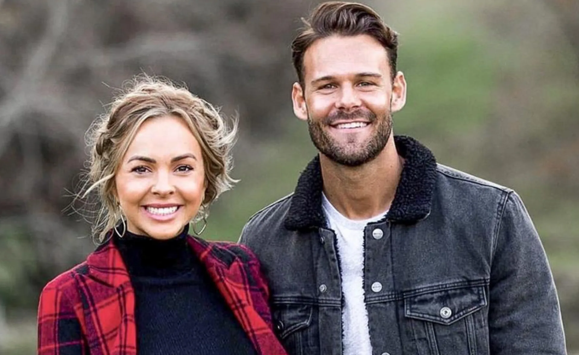 A smiling woman in a red plaid coat and a man in a denim jacket pose outdoors with blurred trees in the background.