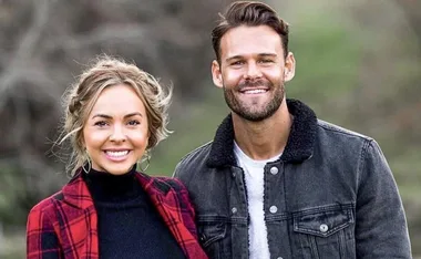 A smiling woman in a red plaid coat and a man in a denim jacket pose outdoors with blurred trees in the background.