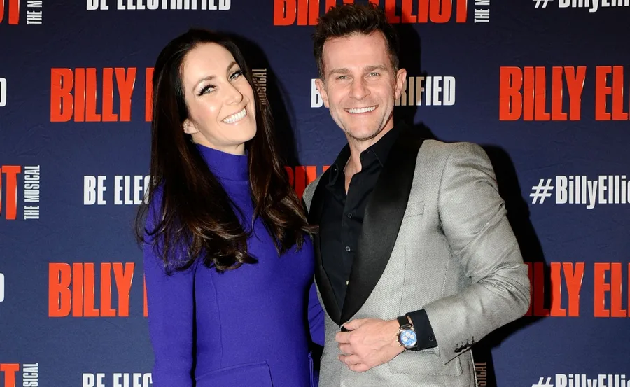 A couple smiling in front of a "Billy Elliot the Musical" backdrop.