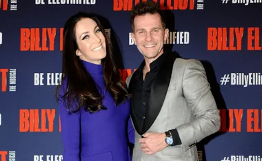 A couple smiling in front of a "Billy Elliot the Musical" backdrop.
