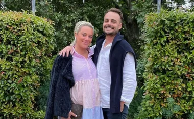 Two people smiling outdoors, standing close together in front of green bushes.