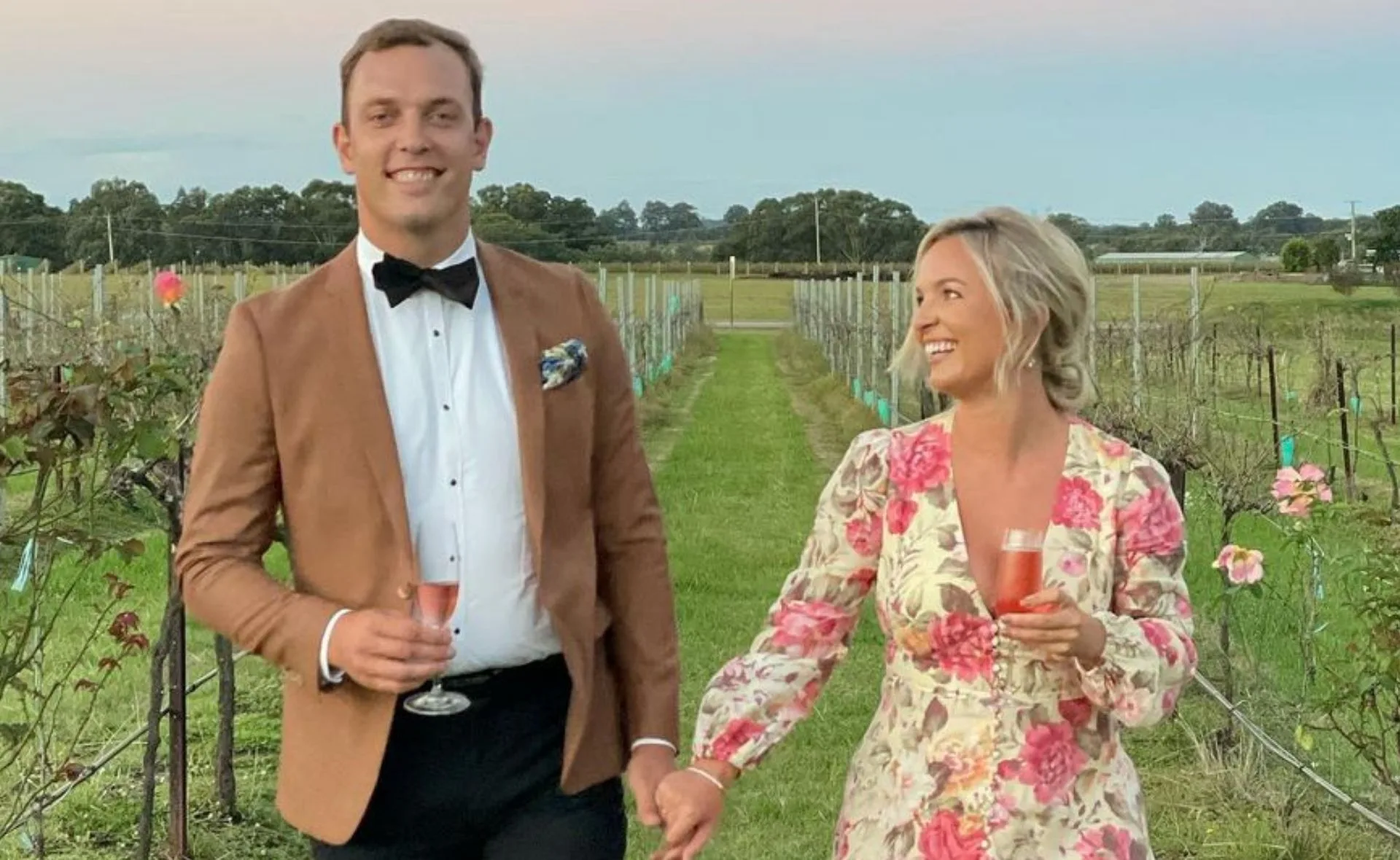 Couple holding hands and smiling, standing in a vineyard, dressed in formal attire.