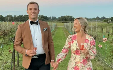 Couple holding hands and smiling, standing in a vineyard, dressed in formal attire.