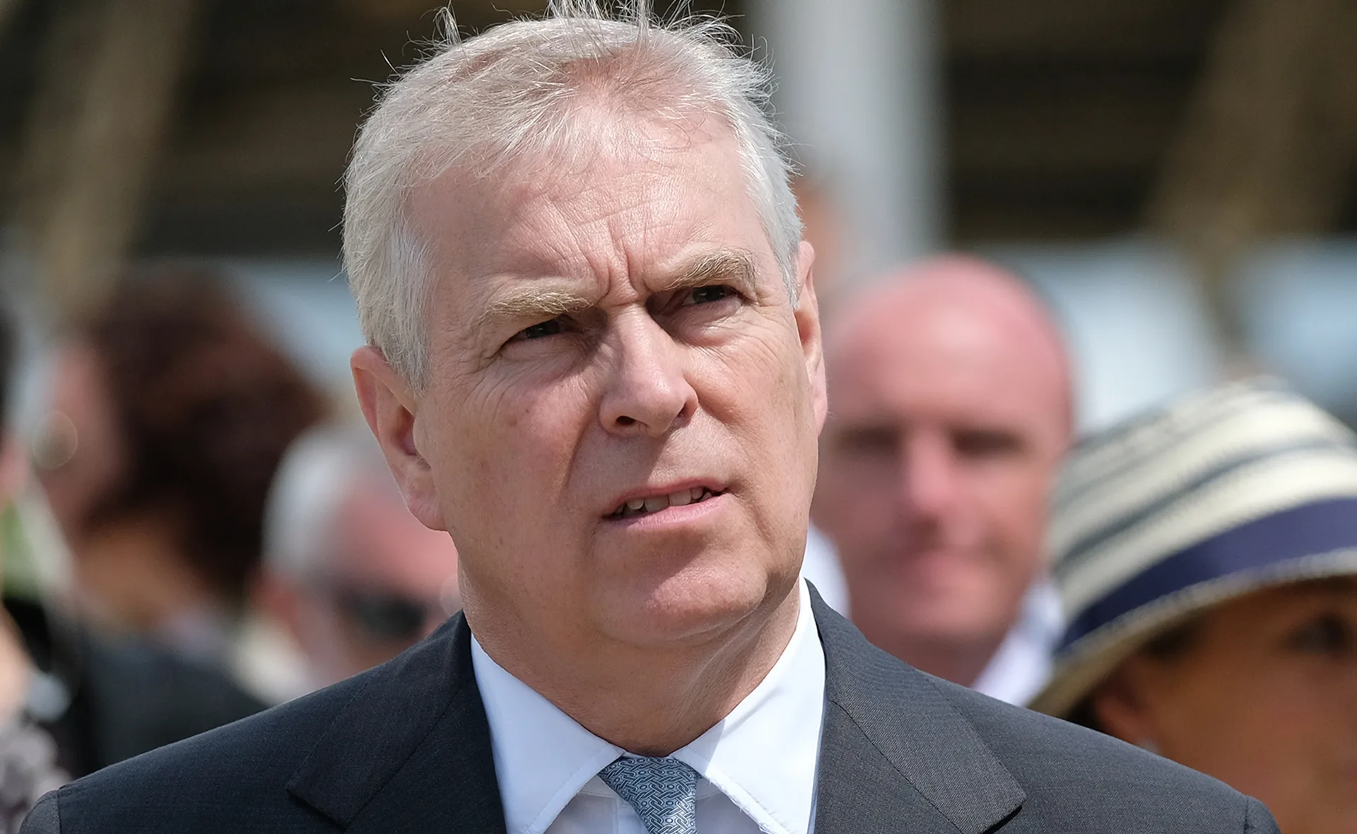A man in a suit and tie with a serious expression, outdoors with a crowd in the background.