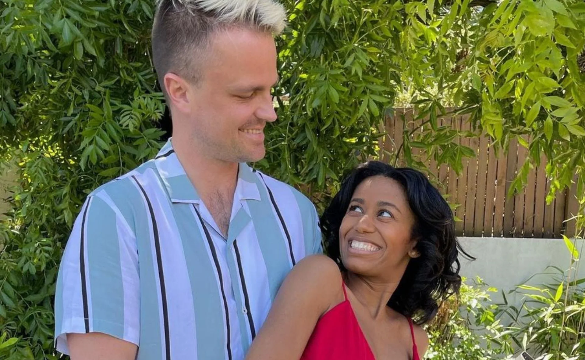 A couple smiling at each other outdoors, surrounded by green foliage.