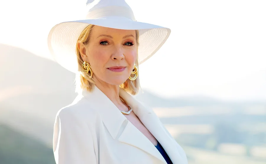 A woman in a white outfit and hat stands outdoors, with a serene landscape background.