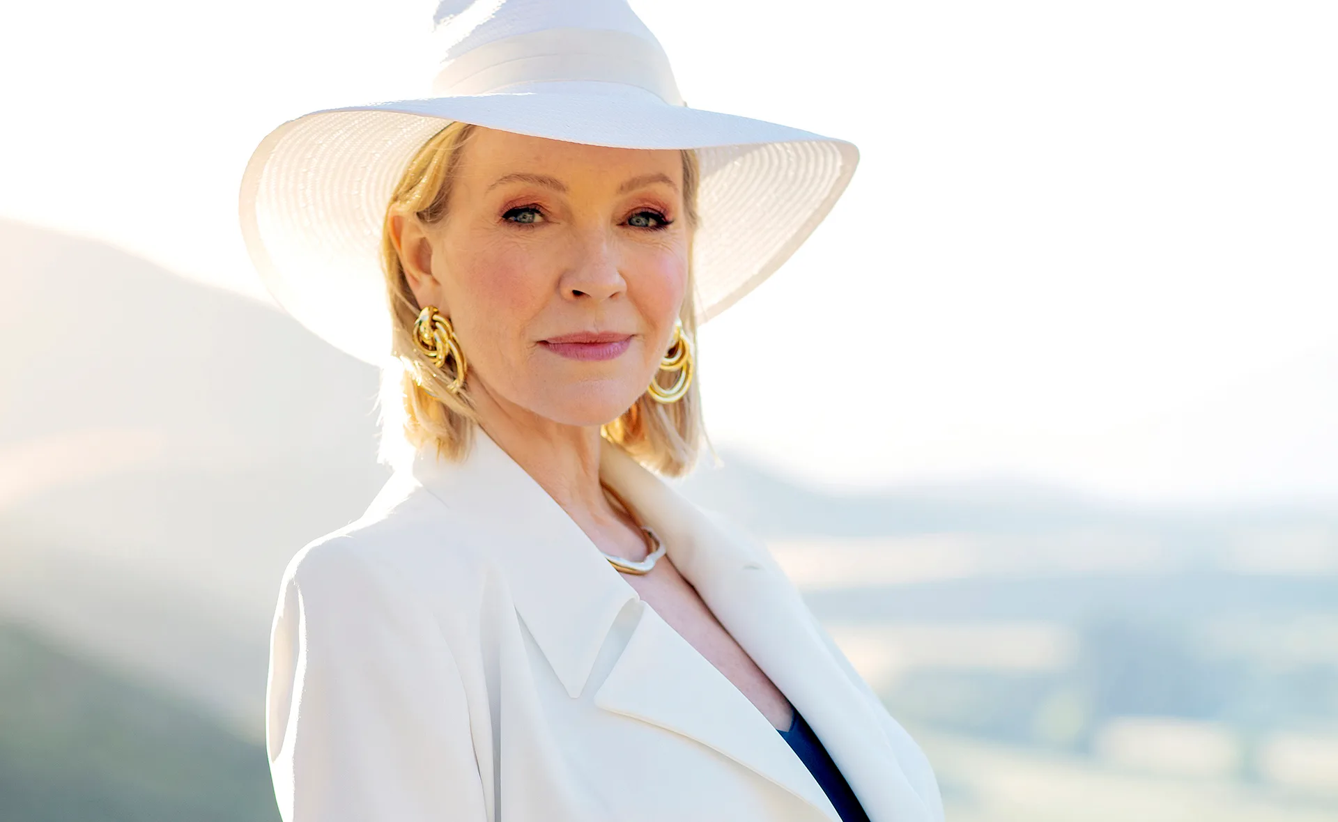 A woman in a white outfit and hat stands outdoors, with a serene landscape background.