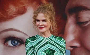 Actress in a green sequin dress with curly hair, smiling, against a backdrop showing a large eye and face.