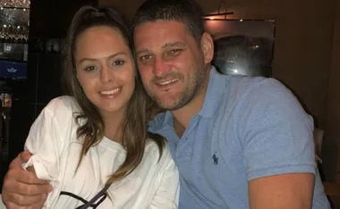 A man and a woman smiling together indoors, the man in a blue shirt hugging the woman in a white shirt.