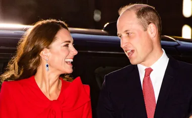 Man and woman smiling at each other, she in red attire with blue earrings, standing near a car at night.