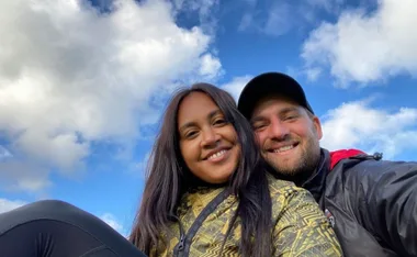 A couple smiling in a selfie with a blue sky and clouds in the background.