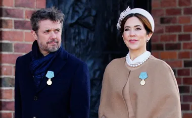 Two elegantly dressed individuals wearing medals are standing outside near a brick wall.