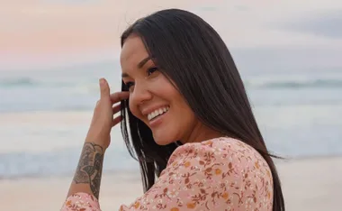 Smiling woman with long hair and tattoo, wearing floral dress at the beach.