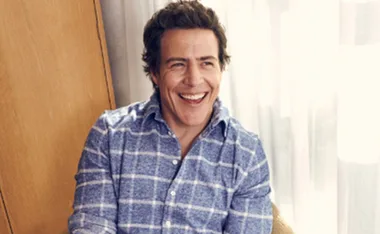 Man in a blue checkered shirt smiling broadly, sitting indoors near a light curtain.