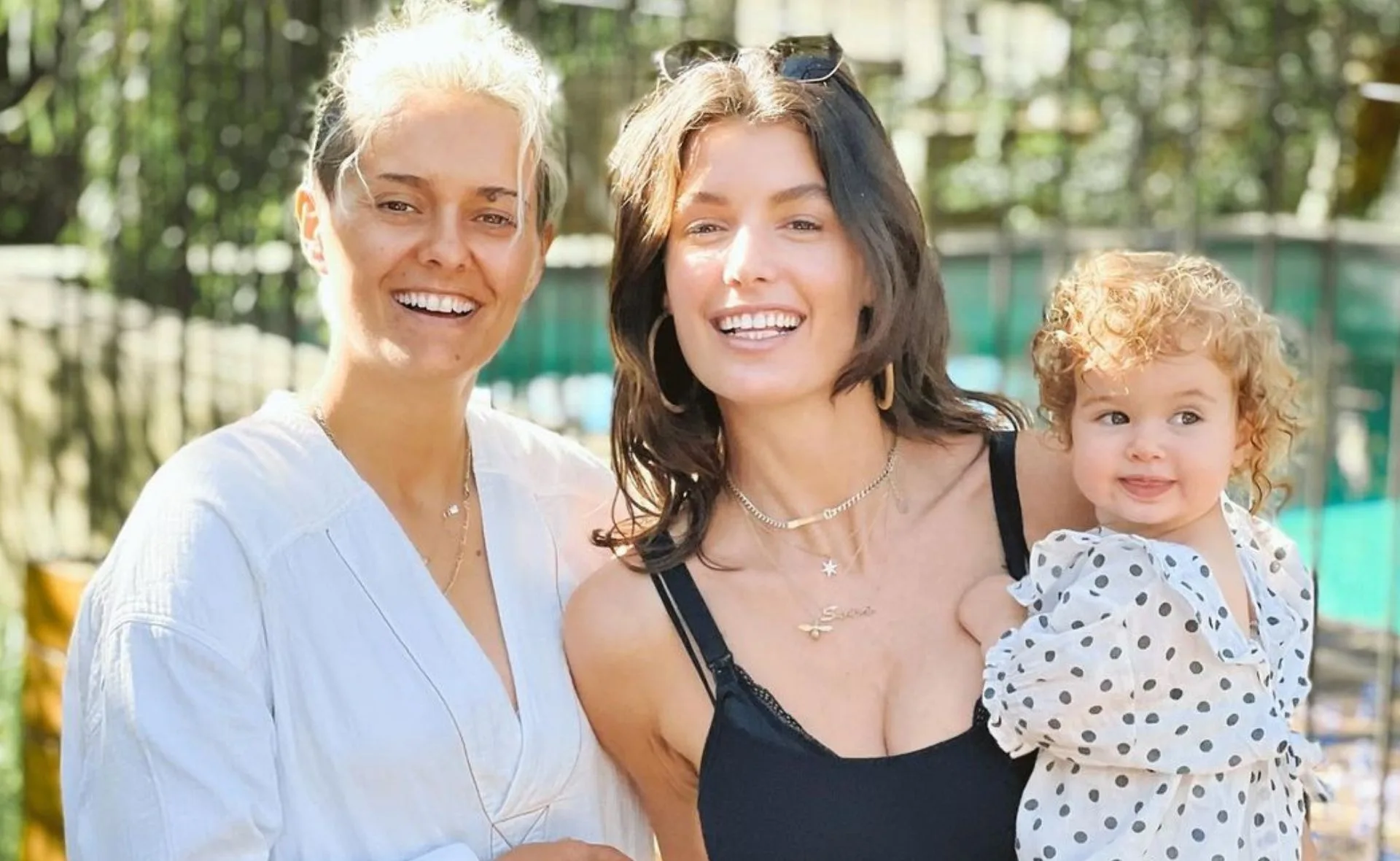 Two smiling adults and a child with curly hair wearing a polka-dot outfit, standing outdoors in sunlight.