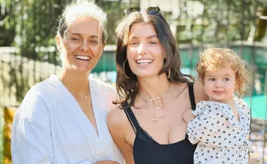 Two smiling adults and a child with curly hair wearing a polka-dot outfit, standing outdoors in sunlight.