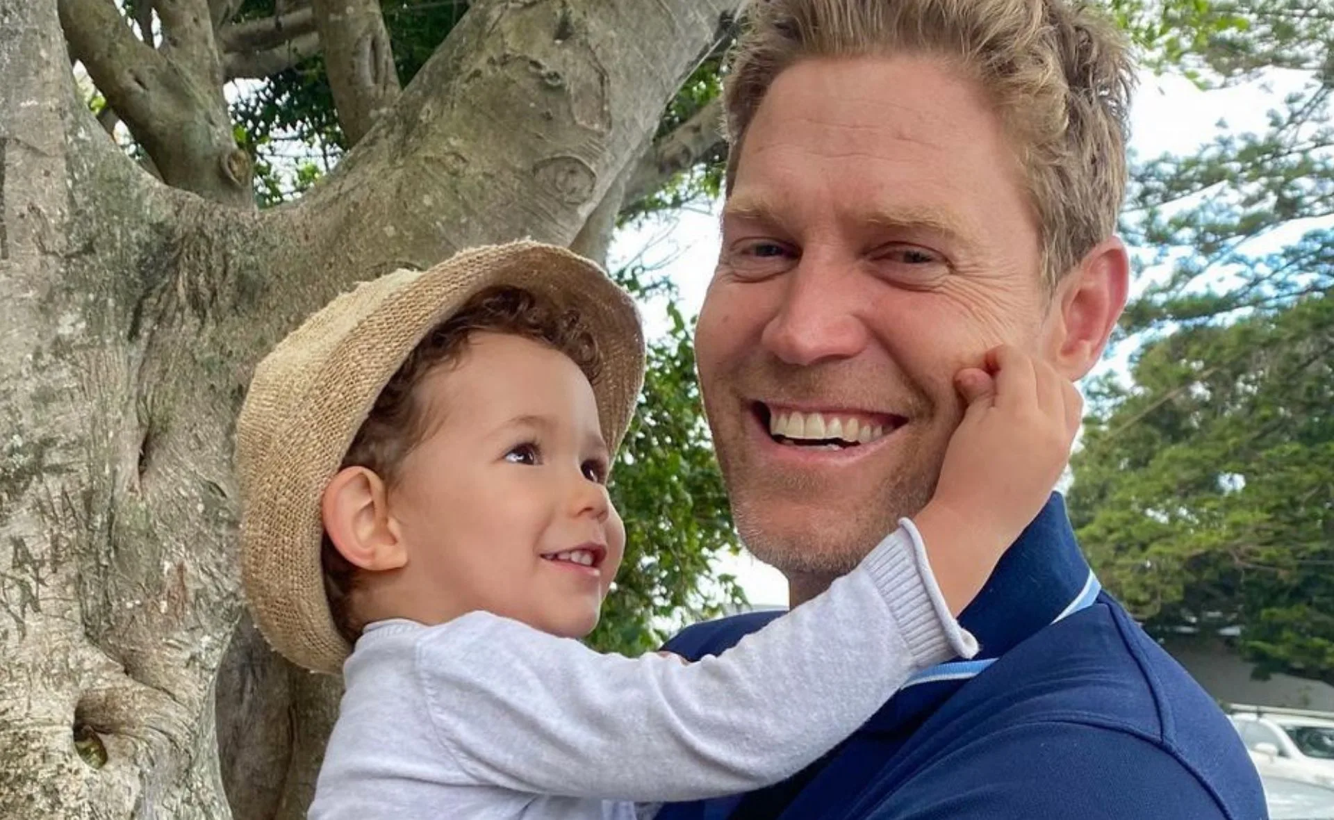 Father holding smiling child in a hat, outdoors near a tree.