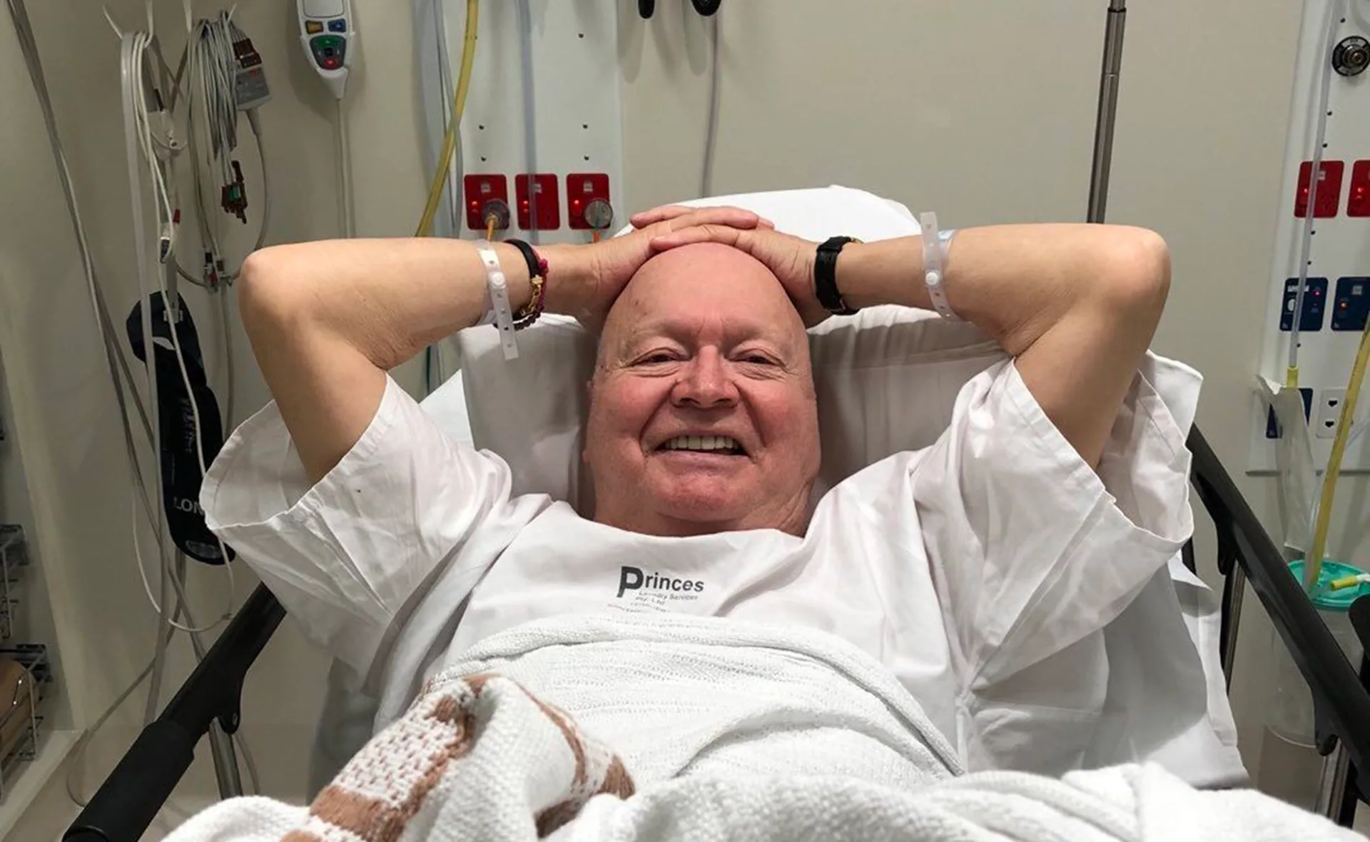 Man smiling in hospital bed with hands behind head, wearing wristbands and a hospital gown.