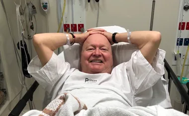 Man smiling in hospital bed with hands behind head, wearing wristbands and a hospital gown.