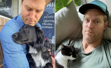Man cuddling with a black dog on the left and holding a black and white cat on the right.