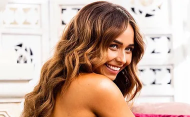 Smiling woman with long, wavy hair, wearing a strapless top, seated in front of a white patterned background.
