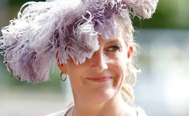 A woman smiling while wearing a hat with large, light purple feathers, with a blurred background.
