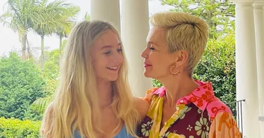 A woman and her daughter smiling at each other on a porch with greenery in the background.