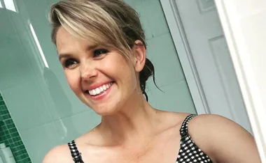 Smiling woman taking a selfie in a bathroom, wearing a checkered top.