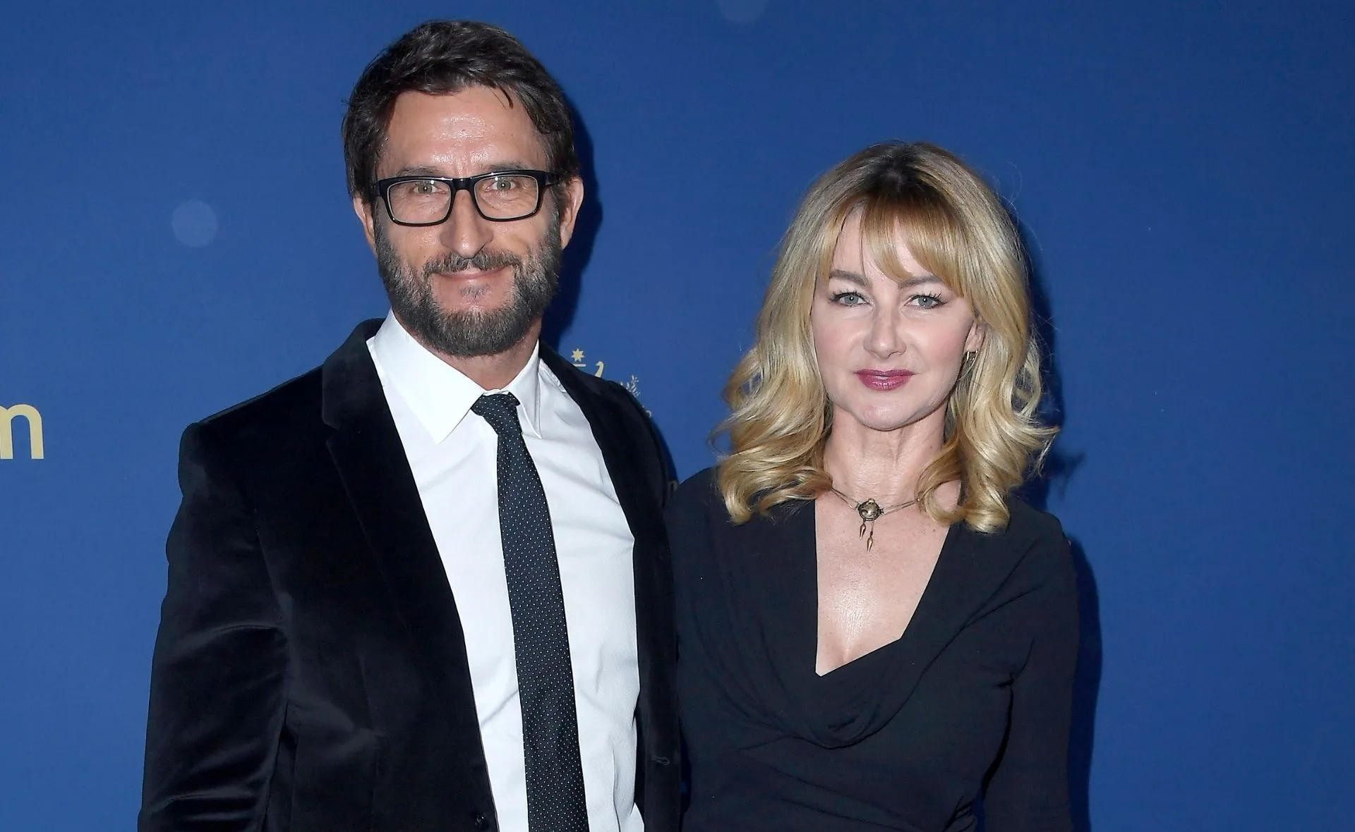 A man in a suit and glasses stands next to a woman in a black dress on a blue background.