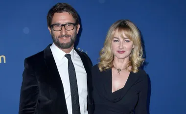 A man in a suit and glasses stands next to a woman in a black dress on a blue background.